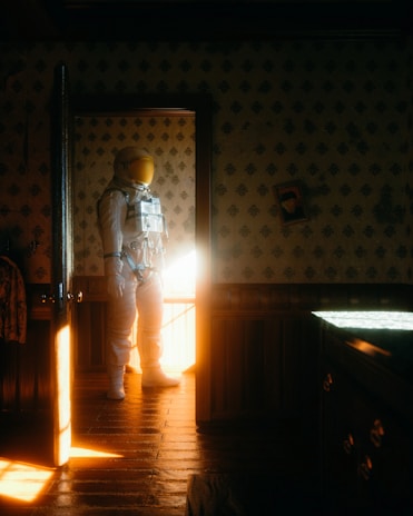 An astronaut wearing a white spacesuit stands in a dimly lit room with patterned wallpaper and wood paneling. Sunlight streams through the open door, casting a warm glow on the floor and the suit. A slightly tilted framed photo hangs on the wall.