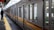 A long passenger train is stationed at an indoor platform with a person standing nearby. The platform has a yellow safety line and there are overhead signs visible.