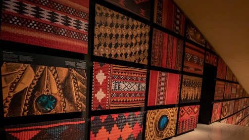 A wall display featuring a variety of traditional woven textile patterns and intricate carvings arranged in a grid formation. The designs include geometric shapes, symmetrical arrangements, and vibrant colors. Some of the sections showcase detailed carvings with circular motifs and turquoise inlays.