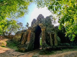 An ancient stone gateway stands amidst a lush green forest, partially covered by creeping vegetation. The structure appears weathered, with intricate carvings and a large central archway. Sunlight filters through the surrounding trees, casting shadows on the path leading through the arch.