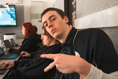 A young person seated in a room with a couple of others, holding up a peace sign with their hand. The environment appears to be a classroom or study area with laptops on the desk. The individuals are focused, possibly engaging in an activity related to computers.