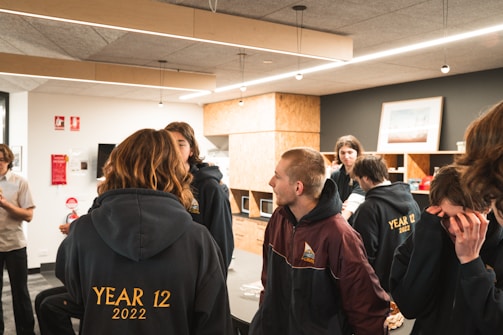 A group of young people are gathered in a modern room, likely a classroom or common area. They are wearing matching year 12 jackets. The room has wooden shelves, a framed picture, and light fixtures hanging from the ceiling.