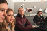 Several young people are sitting in a classroom, appearing focused and content. They are seated at a table with one person using a laptop. The background features posters and papers on a grey wall.