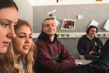 Several young people are sitting in a classroom, appearing focused and content. They are seated at a table with one person using a laptop. The background features posters and papers on a grey wall.