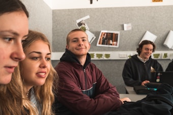 Several young people are sitting in a classroom, appearing focused and content. They are seated at a table with one person using a laptop. The background features posters and papers on a grey wall.