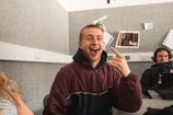 A young man wearing a maroon jacket is sitting at a table, making a playful expression with his tongue out and pointing his index finger. Behind him, another person is sitting at a desk with a laptop. The room has gray walls with some papers pinned on them.