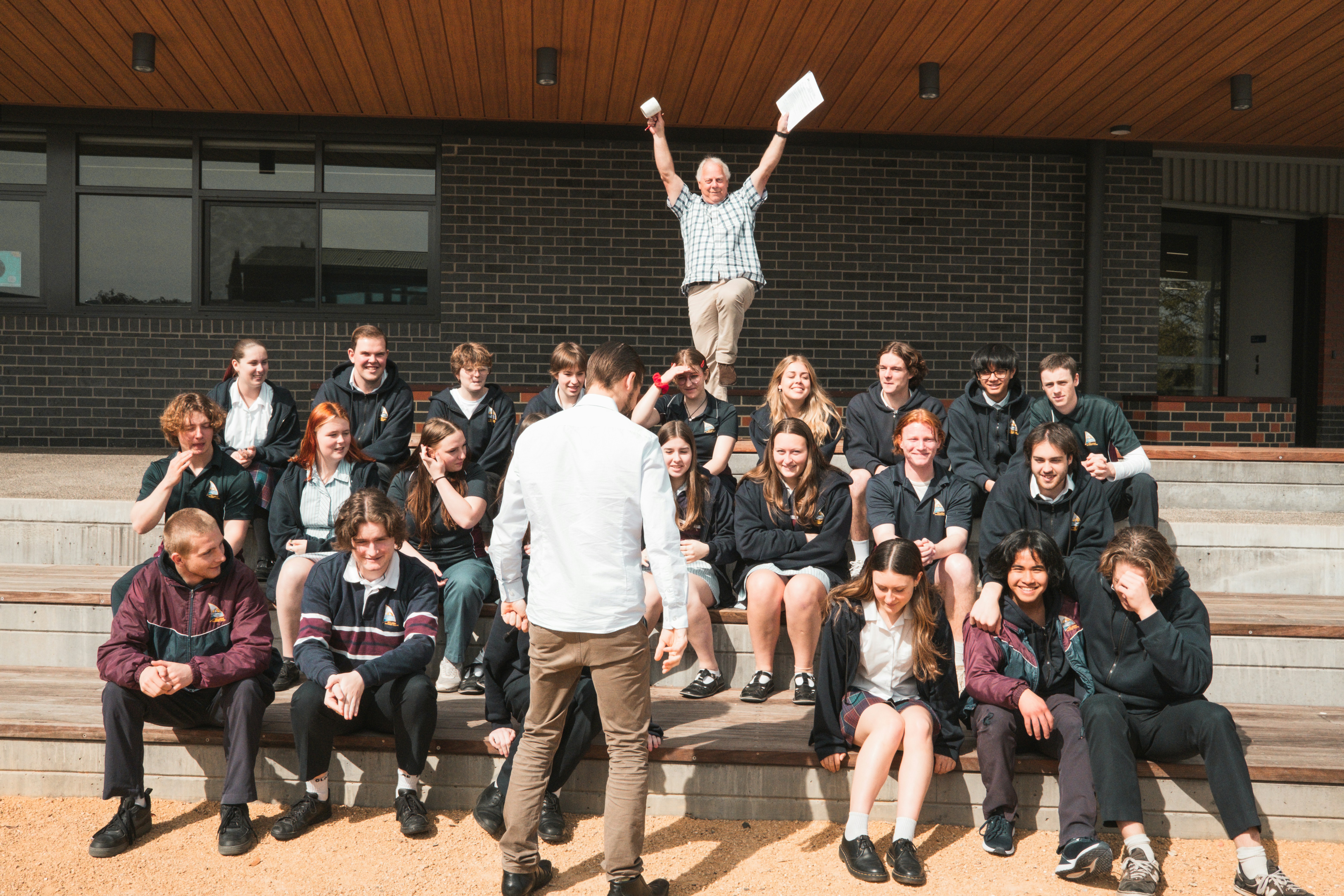 A group of students is seated on outdoor steps, posing for a photo. There is an atmosphere of camaraderie and casual interaction. Above them, an older man, possibly a teacher, energetically raises his arms, holding papers, which adds a sense of celebration or achievement. The background features a modern building with brick walls.