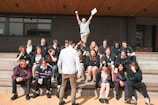 A group of students is seated on outdoor steps, posing for a photo. There is an atmosphere of camaraderie and casual interaction. Above them, an older man, possibly a teacher, energetically raises his arms, holding papers, which adds a sense of celebration or achievement. The background features a modern building with brick walls.