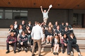 A group of students is seated on outdoor steps, posing for a photo. There is an atmosphere of camaraderie and casual interaction. Above them, an older man, possibly a teacher, energetically raises his arms, holding papers, which adds a sense of celebration or achievement. The background features a modern building with brick walls.