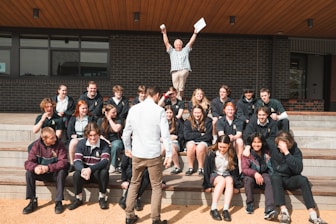 A group of students is seated on outdoor steps, posing for a photo. There is an atmosphere of camaraderie and casual interaction. Above them, an older man, possibly a teacher, energetically raises his arms, holding papers, which adds a sense of celebration or achievement. The background features a modern building with brick walls.