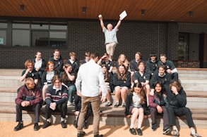 A group of students is seated on outdoor steps, posing for a photo. There is an atmosphere of camaraderie and casual interaction. Above them, an older man, possibly a teacher, energetically raises his arms, holding papers, which adds a sense of celebration or achievement. The background features a modern building with brick walls.
