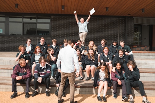 A group of students is seated on outdoor steps, posing for a photo. There is an atmosphere of camaraderie and casual interaction. Above them, an older man, possibly a teacher, energetically raises his arms, holding papers, which adds a sense of celebration or achievement. The background features a modern building with brick walls.