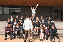 A group of students is seated on outdoor steps, posing for a photo. There is an atmosphere of camaraderie and casual interaction. Above them, an older man, possibly a teacher, energetically raises his arms, holding papers, which adds a sense of celebration or achievement. The background features a modern building with brick walls.