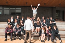 A group of students is seated on outdoor steps, posing for a photo. There is an atmosphere of camaraderie and casual interaction. Above them, an older man, possibly a teacher, energetically raises his arms, holding papers, which adds a sense of celebration or achievement. The background features a modern building with brick walls.