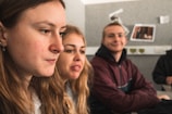 Three people are seated indoors, engaged in conversation. The focus is on a young woman with long brown hair. Two others, one with blonde hair and another with short hair wearing a jacket, are in the background. The setting appears to be a casual or educational environment with a gray wall and some paper items pinned up.