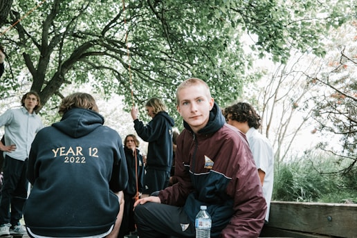 A group of young adults gathered outdoors underneath trees with lush green leaves. One person is seated on a wooden bench wearing a maroon and navy jacket, holding a water bottle, looking directly at the camera. Another has their back to the camera wearing a hoodie that reads 'Year 12 2022', while others engage in conversation nearby.
