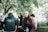 A group of young adults gathered outdoors underneath trees with lush green leaves. One person is seated on a wooden bench wearing a maroon and navy jacket, holding a water bottle, looking directly at the camera. Another has their back to the camera wearing a hoodie that reads 'Year 12 2022', while others engage in conversation nearby.