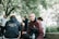 A group of young adults gathered outdoors underneath trees with lush green leaves. One person is seated on a wooden bench wearing a maroon and navy jacket, holding a water bottle, looking directly at the camera. Another has their back to the camera wearing a hoodie that reads 'Year 12 2022', while others engage in conversation nearby.