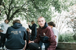 A group of young adults gathered outdoors underneath trees with lush green leaves. One person is seated on a wooden bench wearing a maroon and navy jacket, holding a water bottle, looking directly at the camera. Another has their back to the camera wearing a hoodie that reads 'Year 12 2022', while others engage in conversation nearby.