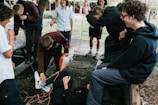 A group of young people is gathered outdoors, involved in a playful interaction. One person is lying on the ground wrapped in orange rope, while others are watching and laughing. The setting appears to be a park or garden with trees and benches.