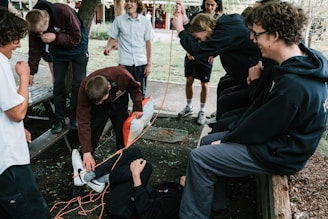 A group of young people is gathered outdoors, involved in a playful interaction. One person is lying on the ground wrapped in orange rope, while others are watching and laughing. The setting appears to be a park or garden with trees and benches.