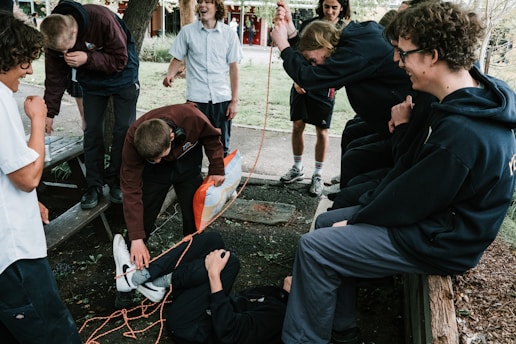 A group of young people is gathered outdoors, involved in a playful interaction. One person is lying on the ground wrapped in orange rope, while others are watching and laughing. The setting appears to be a park or garden with trees and benches.