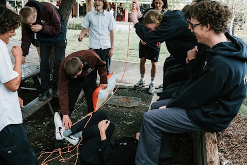 A group of young people is gathered outdoors, involved in a playful interaction. One person is lying on the ground wrapped in orange rope, while others are watching and laughing. The setting appears to be a park or garden with trees and benches.