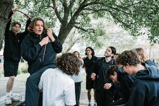 A group of young people gathered outdoors under a tree. Some individuals are wearing dark hoodies, while others are dressed casually. The atmosphere appears lively and playful, with one person holding onto a rope tied to a tree branch. The presence of trees and natural surroundings suggests a park or outdoor recreational area.