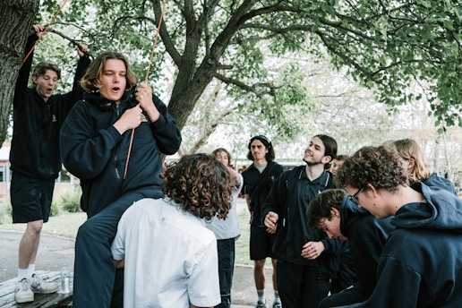 A group of young people gathered outdoors under a tree. Some individuals are wearing dark hoodies, while others are dressed casually. The atmosphere appears lively and playful, with one person holding onto a rope tied to a tree branch. The presence of trees and natural surroundings suggests a park or outdoor recreational area.