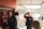 Several students are in a school hallway, some interacting with lockers. The scene is outdoors with an overhead canopy and a sidewalk beside a brick building. A group of students is engaging in casual conversation or activity.