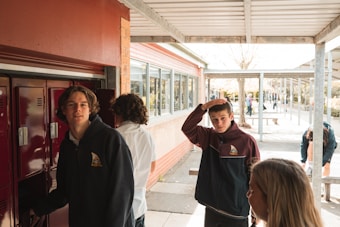 Several students are in a school hallway, some interacting with lockers. The scene is outdoors with an overhead canopy and a sidewalk beside a brick building. A group of students is engaging in casual conversation or activity.