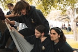 A group of young people are gathered around an outdoor setting with trees and some sort of overhead shelter. Three individuals are the focus, dressed in dark hoodies and interacting playfully with fabric or a hammock. Their expressions suggest amusement and camaraderie. Others in the background appear to be engaged in conversations or walking, hinting at a lively atmosphere.