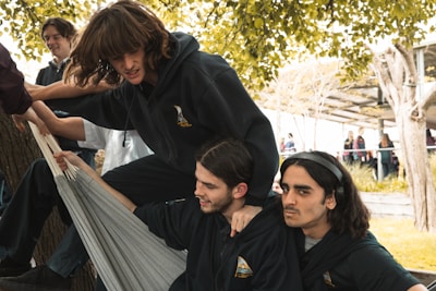 A group of young people are gathered around an outdoor setting with trees and some sort of overhead shelter. Three individuals are the focus, dressed in dark hoodies and interacting playfully with fabric or a hammock. Their expressions suggest amusement and camaraderie. Others in the background appear to be engaged in conversations or walking, hinting at a lively atmosphere.