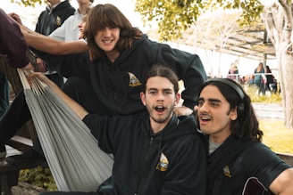 Several people are gathered outdoors, with three individuals prominently seated. They are smiling and appear relaxed, wearing matching dark hoodies with a logo, seated on what seems to be a hammock.