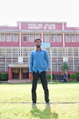 A man stands on a grassy lawn in front of a multi-story building with red brick walls and white decorative railings. The building is identified as Kirori Mal College, University of Delhi, and features large windows lined up symmetrically. A group of people can be seen in the background near the entrance.