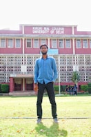 A man stands on a grassy lawn in front of a multi-story building with red brick walls and white decorative railings. The building is identified as Kirori Mal College, University of Delhi, and features large windows lined up symmetrically. A group of people can be seen in the background near the entrance.