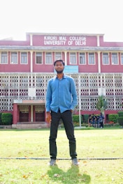 A man stands on a grassy lawn in front of a multi-story building with red brick walls and white decorative railings. The building is identified as Kirori Mal College, University of Delhi, and features large windows lined up symmetrically. A group of people can be seen in the background near the entrance.