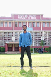 A man stands on a grassy lawn in front of a multi-story building with red brick walls and white decorative railings. The building is identified as Kirori Mal College, University of Delhi, and features large windows lined up symmetrically. A group of people can be seen in the background near the entrance.