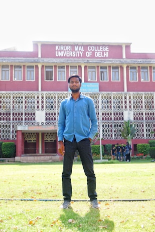A man stands on a grassy lawn in front of a multi-story building with red brick walls and white decorative railings. The building is identified as Kirori Mal College, University of Delhi, and features large windows lined up symmetrically. A group of people can be seen in the background near the entrance.