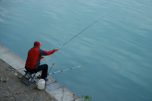 A person wearing a red jacket and cap is fishing while sitting on a small chair by the waterside. The fishing rod is extended over the calm water, and there's a white container beside the individual, suggesting it's being used as a bait or equipment holder. The surrounding area has a stone and cement pavement.