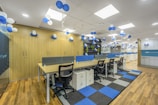 A modern office space decorated with clusters of blue and white balloons hanging from the ceiling. The room features rows of desks with ergonomic chairs, storage cabinets, and blue accents. The floor has a pattern of wooden and carpeted tiles, and the walls include shelving with plants and additional decor.
