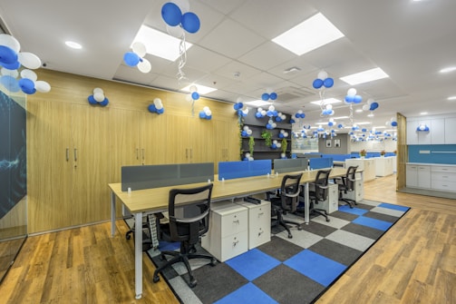 A modern office space decorated with clusters of blue and white balloons hanging from the ceiling. The room features rows of desks with ergonomic chairs, storage cabinets, and blue accents. The floor has a pattern of wooden and carpeted tiles, and the walls include shelving with plants and additional decor.
