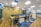 A modern office space decorated with clusters of blue and white balloons hanging from the ceiling. The room features rows of desks with ergonomic chairs, storage cabinets, and blue accents. The floor has a pattern of wooden and carpeted tiles, and the walls include shelving with plants and additional decor.