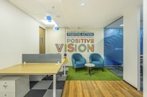 A modern office space featuring a wooden desk with a partition on the left side. Adjacent to it is a cozy seating area with two teal armchairs and a small round table on a green rug. Behind the seating area, there is a wall with an inspirational quote in colorful text. The floor combines wooden and carpeted sections, and the room is well-lit with overhead lighting and natural light from windows.