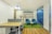 A modern office space featuring a wooden desk with a partition on the left side. Adjacent to it is a cozy seating area with two teal armchairs and a small round table on a green rug. Behind the seating area, there is a wall with an inspirational quote in colorful text. The floor combines wooden and carpeted sections, and the room is well-lit with overhead lighting and natural light from windows.