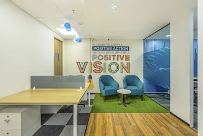 A modern office space featuring a wooden desk with a partition on the left side. Adjacent to it is a cozy seating area with two teal armchairs and a small round table on a green rug. Behind the seating area, there is a wall with an inspirational quote in colorful text. The floor combines wooden and carpeted sections, and the room is well-lit with overhead lighting and natural light from windows.