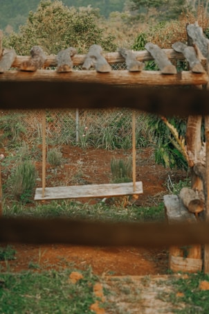 A rustic wooden swing hangs in a lush garden setting, surrounded by greenery and a backdrop of trees. The earth-toned landscape and natural wood elements create a serene and earthy atmosphere.