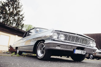 A classic silver car is parked on a paved surface with a low-angle view highlighting its front grille and headlights. The scene includes a backdrop of a building and some trees, adding a rustic atmosphere.