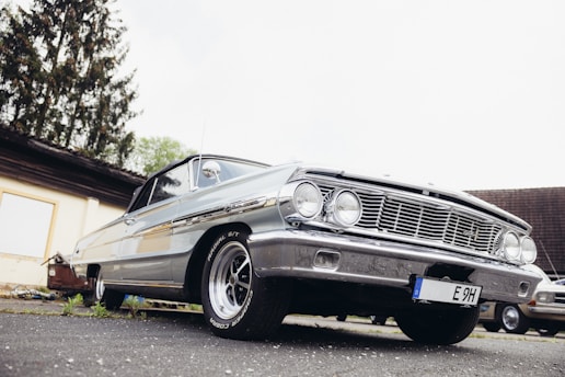 A classic silver car is parked on a paved surface with a low-angle view highlighting its front grille and headlights. The scene includes a backdrop of a building and some trees, adding a rustic atmosphere.