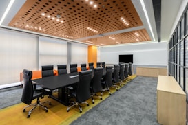 A modern conference room with a long black table surrounded by black leather swivel chairs. The room features a stylish ceiling with circular wooden patterns and recessed lighting. There is a large flat-screen TV mounted on the wall, and abundant natural light comes through large glass windows. The carpet is gray with a yellow strip near the table, and the walls are light-colored with a section in vibrant orange.